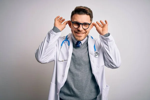 青い目の若い医者の男は 孤立した背景の上に医療用コートと聴診器を身に着けています指で耳を引く笑顔 面白いジェスチャー オーディション問題 — ストック写真