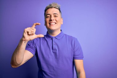 İzole edilmiş arka plan üzerinde sıradan mor tişört giyen genç, modern bir adam gülümseyen ve kendine güvenen el hareketleriyle küçük boy işareti yapan parmaklarıyla ve kamerayla. Ölçü kavramı.