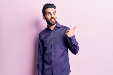 Sakallı, sıradan tişörtlü, mutlu yüzlü, gülümseyen ve baş parmağıyla yan tarafı gösteren yakışıklı bir genç.. 