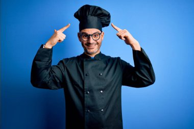 Genç, yakışıklı aşçı üniformalı ve şapkalı bir adam izole edilmiş mavi arka planda gülümsüyor iki eliyle başını işaret ediyor, harika bir fikir ya da düşünce, iyi bir hafıza.