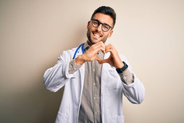 Gözlük takan genç doktor, tıbbi beyaz bornoz ve izole edilmiş arka planda steteskop takan, aşkla gülümseyen, elleriyle kalp sembolü yapan genç bir adam. Romantik konsept.