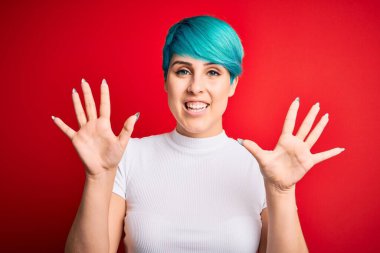 Mavi saçlı, kırmızı arka planda günlük tişört giyen genç ve güzel bir kadın on numarayı gösterirken kendinden emin ve mutlu bir şekilde gülümsüyor..