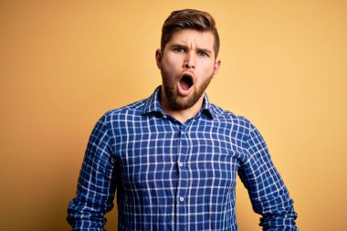 Sarışın, sakallı ve mavi gözlü genç bir işadamı sarı arka planda tişört giyiyor. Şok olmuş yüz ifadesiyle şüpheci ve alaycı görünüyor.