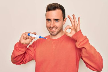 Mavi gözlü genç adam dijital termometre kullanarak sıcaklığı ölçüyor. Parmaklarıyla işaret veriyor, gülümsüyor ve mükemmel bir sembol oluşturuyor.