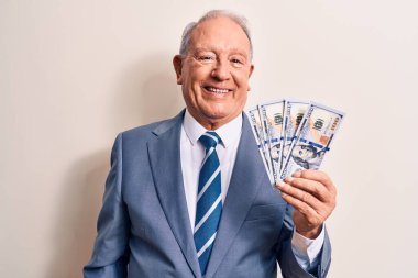 Kıdemli, gri saçlı, takım elbiseli bir iş adamı elinde bir deste dolar banknotlarla mutlu ve pozitif görünüyor ve gülümsüyor dişlerini gösteren bir gülümsemeyle.