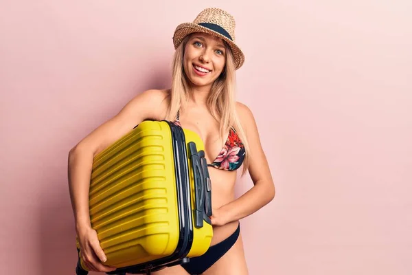Young beautiful blonde woman wearing bikini and hat holding cabin bag looking positive and happy standing and smiling with a confident smile showing teeth