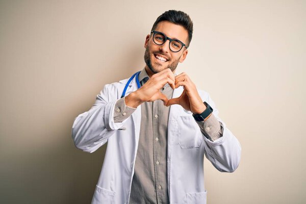 Young doctor man wearing glasses, medical white robe and stethoscope over isolated background smiling in love doing heart symbol shape with hands. Romantic concept.