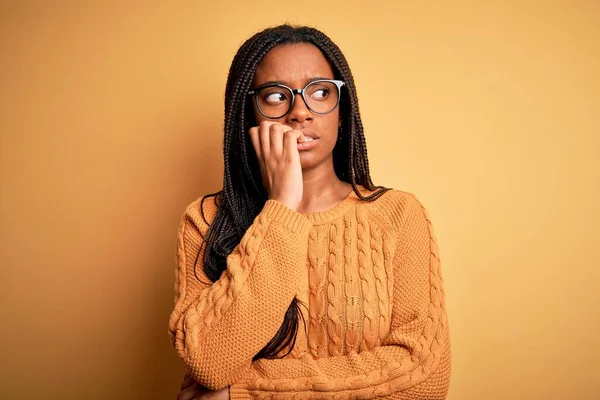 黄色の背景に眼鏡とカジュアルなセーターを身に着けている若いアフリカ系アメリカ人のスマート女性は 口の爪をかむ上で手で強調し 緊張している 不安の問題 — ストック写真