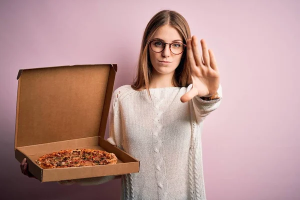 漂亮的红头发女人拿着装意大利新鲜披萨的分娩盒 粉红的背景 张开手做停止手势 严肃而自信的表情 防守的姿态 — 图库照片