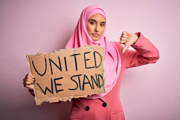 Young activist woman wearing pink muslim hijab holding banner with united we stand message with angry face, negative sign showing dislike with thumbs down, rejection concept