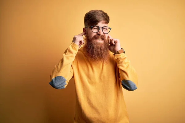 Handsome Irish redhead man with beard wearing glasses over yellow isolated background covering ears with fingers with annoyed expression for the noise of loud music. Deaf concept.