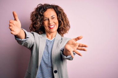 Soyutlanmış pembe arka planda zarif bir ceket giymiş orta yaşlı güzel bir iş kadını kameraya sarılmak için gülümseyerek bakıyor. Mutluluğu kucaklayan neşeli bir ifade..