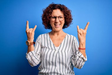 Orta yaşlı güzel kıvırcık saçlı kadın izole arka planda sıradan çizgili tişört giyiyor çılgın bir ifadeyle elleri havada rock sembolü yapıyor. Müzik yıldızı. Ağır konsept.