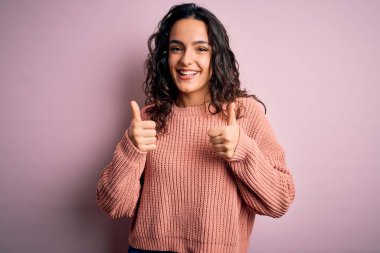 Kıvırcık saçlı genç bir kadın pembe arka plan işareti üzerine günlük süveter giyiyor. Elleriyle olumlu jest yapıyor. Gülümsüyor ve mutlu. Neşeli ifade ve kazanan jest.