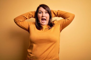 Güzel esmer ve cüsseli bir kadın izole edilmiş sarı arka planda günlük süveter giyiyor. Deli ve elleri kafasında korkmuş, ağzı açık şoktan korkmuş ve şaşırmış.