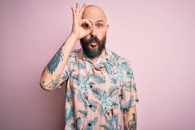 Sakallı, dövmeli, pembe arka planda sıradan bir çiçek gömleği giyen yakışıklı, şaşırmış yüz ifadesiyle şaşırmış, parmaklarından bakan bir adam. İnançsız ifade.