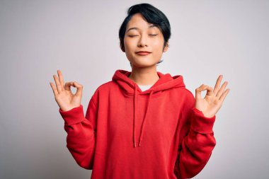 Genç, güzel Asyalı bir kız beyaz arka planda kapüşonlu sıradan bir sweatshirt giyiyor. Gözleri kapalı gülümsüyor ve parmaklarıyla meditasyon hareketi yapıyor. Yoga konsepti.