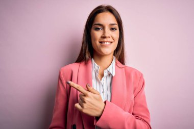 Genç ve güzel esmer kadın ceketini giyiyor pembe arka planda neşeli bir şekilde duruyor. Yüzünde mutlu ve doğal bir ifadeyle elleri ve parmaklarıyla yüzünü gösteren bir gülümseme var.