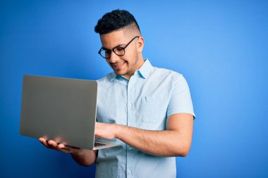 Gözlüklü genç yakışıklı iş adamı mavi arka planda dizüstü bilgisayar kullanıyor. Mutlu bir yüzü var. Kendine güvenen bir gülümsemeyle dişlerini gösteriyor.