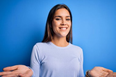Genç, güzel, esmer bir kadın mavi arka planda dikilmiş neşeli gülümsüyor, dostça karşılıyor, olumlu ve kendinden emin selamlıyor.