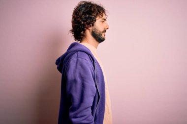 Sakallı, pembe arka plan üzerine sıradan bir kazak giyen, rahat bir görünüm takınan, doğal yüzlü ve kendinden emin gülümseyen genç yakışıklı bir adam..