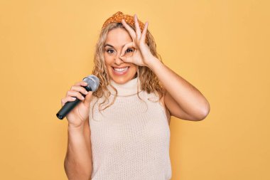Sarı arka planda mikrofon kullanarak şarkı söyleyen güzel sarışın kadın gülümsüyor. Tamam işareti yaparken parmaklarının arasından bakıyor.