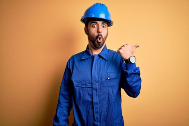 Sakallı tamirci, mavi üniformalı ve sarı arka planda kasklı. El parmağı yana dönük, ağzı açık, şaşırmış bir halde..