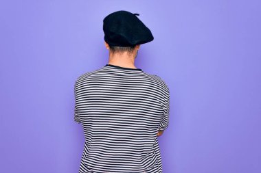 Mor arka plan üzerinde çizgili tişört ve Fransız beresi giyen, kollarını kavuşturup arkaya bakan yakışıklı bir adam.