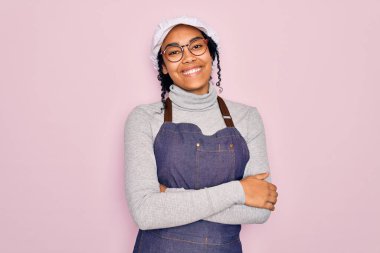 Genç Afro-Amerikan kıvırcık fırıncı kadın pembe arka planda önlük ve şapka takıyor ve kollarını kavuşturup kameraya bakıyor. Pozitif kişi.