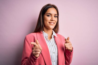 İzole edilmiş pembe arka planda duran ceketli güzel esmer kadın mutlu ve komik yüzlü parmaklarını kameraya doğrultuyor. İyi enerji ve titreşimler.