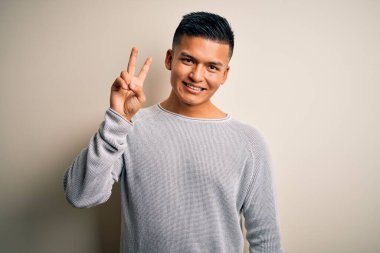 İzole edilmiş beyaz arka planda dikilen gündelik süveter giyen genç ve yakışıklı Latin adam iki numarayı gösterirken kendinden emin ve mutlu bir şekilde gülümsüyor..