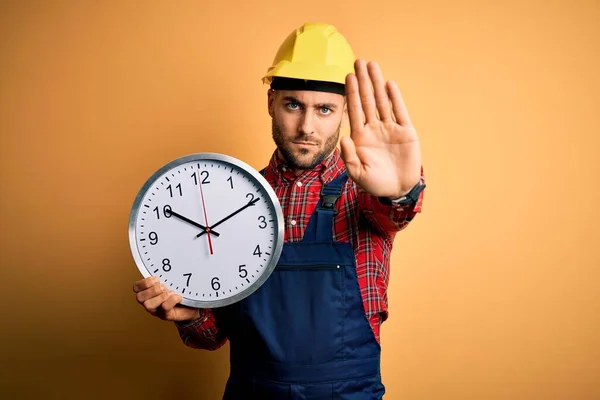 Junger Bauarbeiter Mit Schutzhelm Hält Große Uhr Über Gelbem Hintergrund — Stockfoto