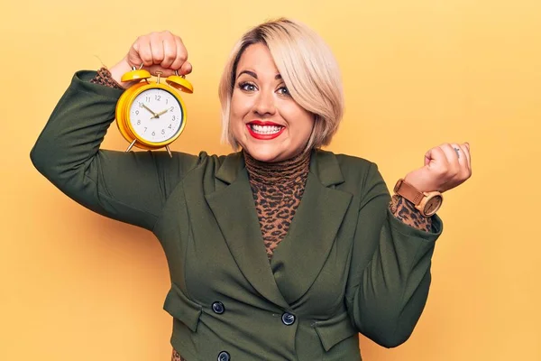Young beautiful plus size blonde woman holding vintage alarm clock over yellow background screaming proud, celebrating victory and success very excited with raised arm