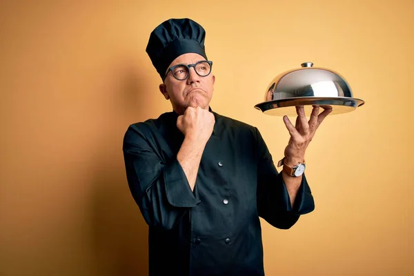 Middle age handsome grey-haired waiter man wearing cooker uniform and hat holding tray with hand on chin thinking about question, pensive expression. Smiling with thoughtful face. Doubt concept.