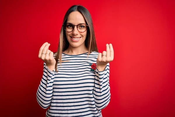 Young beautiful blonde woman with blue eyes wearing glasses standing over red background doing money gesture with hands, asking for salary payment, millionaire business