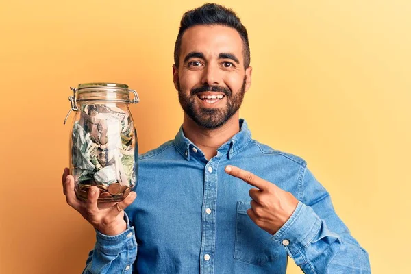 Молодий Іспаномовний Чоловік Тримає Банку Заощадженнями Усміхнений Щасливий Вказує Рукою — стокове фото