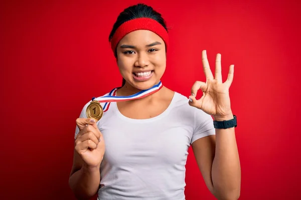 Young asian champion girl winning medal standing over isolated red background doing ok sign with fingers, excellent symbol