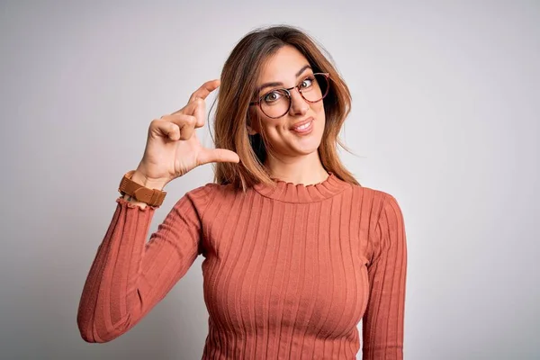 Young beautiful brunette woman wearing casual sweater and glasses over white background smiling and confident gesturing with hand doing small size sign with fingers looking and the camera. Measure concept.
