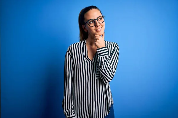 Beautiful blonde woman with blue eyes wearing striped shirt and glasses over blue background with hand on chin thinking about question, pensive expression. Smiling with thoughtful face. Doubt concept.