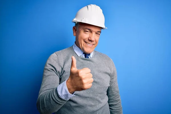 中年英俊 白发苍苍的工程师 头戴安全帽 身披蓝色背景 手举着快乐的大拇指 在镜头前看着成功的表情 — 图库照片