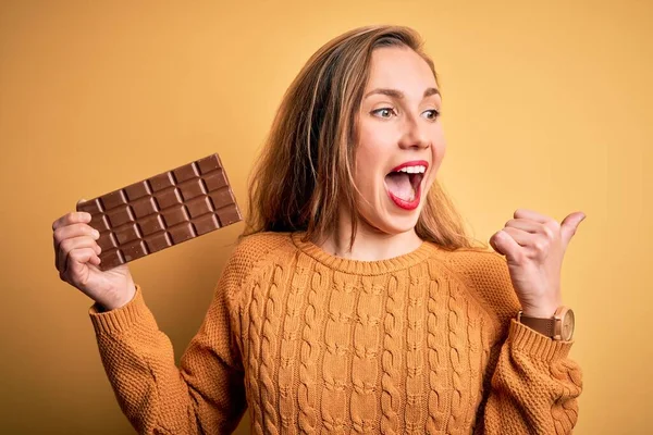 若いです美しいですブロンド女性保持チョコレートバー立っています上の隔離された黄色の背景ポインティングとショーとともに親指でサイドアップとともに幸せな顔笑顔 — ストック写真