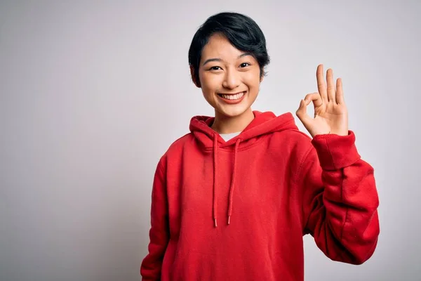 白い背景の上にパーカーを着た若い美しいアジアの女の子は 手と指でOkサインをして積極的に笑顔でカジュアルスウェットシャツを着ています 成功した表現 — ストック写真