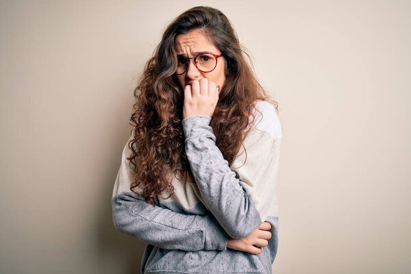 Young beautiful woman with curly hair wearing sweater and glasses over white background looking stressed and nervous with hands on mouth biting nails. Anxiety problem.