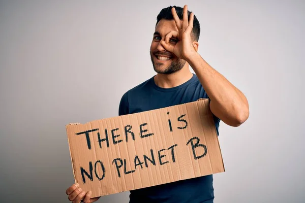 Young handsome activist man protesting for the enviroment hoding cardboard with happy face smiling doing ok sign with hand on eye looking through fingers