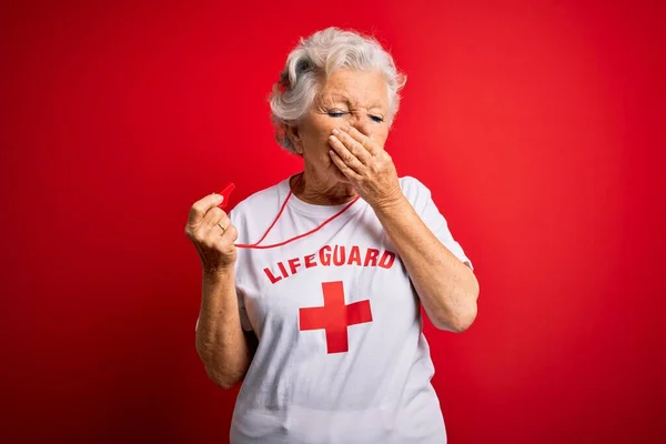 年长美丽的灰白头发的救生员女士穿着印有红十字会标志的T恤衫 用哨子嗅出一股难闻 无法忍受的气味 用手指捂住鼻子屏住呼吸 — 图库照片