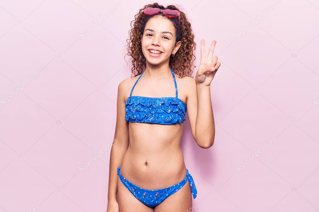 Beautiful kid girl with curly hair wearing bikini and sunglasses showing and pointing up with fingers number two while smiling confident and happy. 