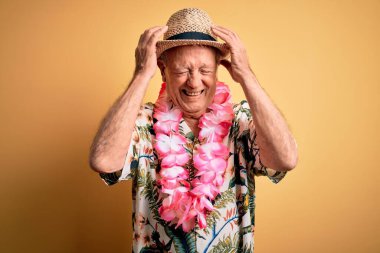 Beyaz saçlı, yaz şapkası takan ve sarı arka planda Hawaii kolyesi takan çaresiz ve stresli yaşlı bir adam. Ağrı ve migren yüzünden. Ellerini başının üstüne koy..