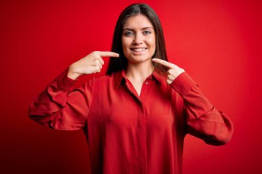 Mavi gözlü genç bir kadın kırmızı arka planda dikilmiş neşeli gülüyor ve parmaklarıyla ve ağzıyla işaret ediyor. Diş sağlığı konsepti.