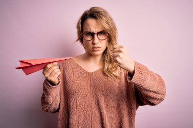 Genç, sarışın, gözlüklü, pembe arka planda kağıt uçak tutan bir kadın. Sinirli ve sinirli bir şekilde bağırıyor, çıldırıyor ve el kaldırarak bağırıyor.