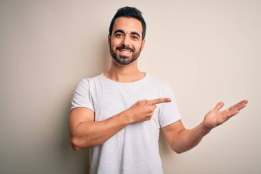 Beyaz arka plan üzerinde duran sıradan tişörtlü, sakallı genç yakışıklı adam kameraya hayret ediyor ve gülümsüyor..